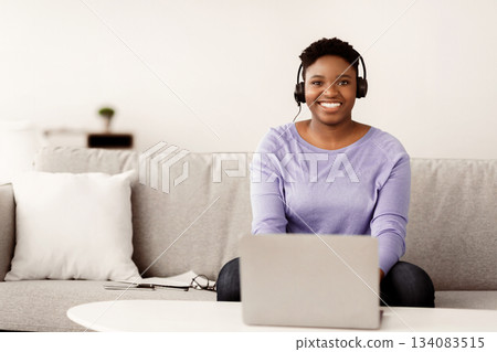 Portrait of smiling female customer service operator in headset posing at workplace, looking at camera, portrait of body positive hotline manager sitting on sofa with laptop in home office, copy space Portrait of smiling female customer service operator in headset posing at workplace, looking at camera, portrait of body positive hotline manager sitting on sofa with laptop in home office, copy space 134083515