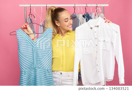 Portrait of young smiling lady standing in clothes shop choosing stylish outfit, holding hangers with dress and shirt in both hands, deciding what to wear, clothing rail in the background Portrait of young smiling lady standing in clothes shop choosing stylish outfit, holding hangers with dress and shirt in both hands, deciding what to wear, clothing rail in the background 134083521