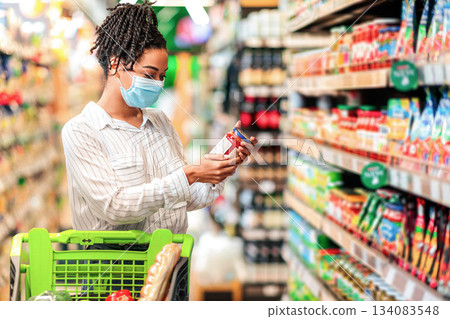 Black Woman Checking Expiration Date On Food Product Doing Grocery Shopping In Supermarket Groceries Store, Standing In Aisle With Shop Trolley. Lady Buyer Choosing Healthy Food Concept Black Woman Checking Expiration Date On Food Product Doing Grocery Shopping In Supermarket Groceries Store, Standing In Aisle With Shop Trolley. Lady Buyer Choosing Healthy Food Concept 134083548