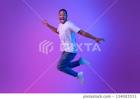 Happy Excited Black Man Jumping With Opened Mouth In Neon Light, Emotional African American Guy In White T-Shirt Having Fun Over Purple Studio Background, Full-Length Shot With Copy Space 134083551