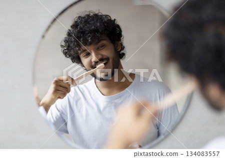 Toothcare routine concept. Young indian guy cleaning teeth in the morning using toothbrush and toothpaste, standing near round mirror in bathroom 134083557