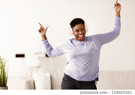 Favourite Playlist Concept. Excited large build african american woman in pink wireless headset listening to music, dancing, resting at home and looking at camera. Young black chubby female having fun Favourite Playlist Concept. Excited large build african american woman in pink wireless headset listening to music, dancing, resting at home and looking at camera. Young black chubby female having fun 134083562