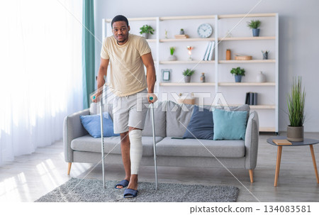Young black guy with plastered leg walking with crutches, suffering from pain at home, copy space. Full length portrait of millennial Afro man having fractured limb. Health care concept Young black guy with plastered leg walking with crutches, suffering from pain at home, copy space. Full length portrait of millennial Afro man having fractured limb. Health care concept 134083581