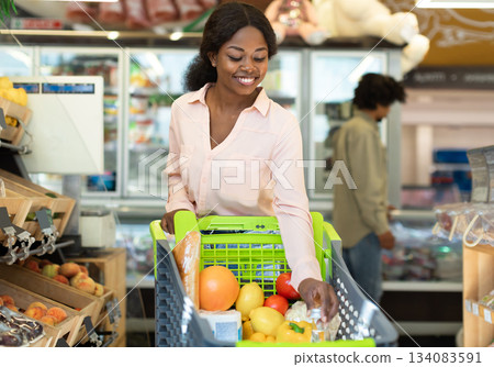 Happy Black Female Buyer Walking With Shop Cart Full Of Food Doing Grocery Shopping In Modern Supermarket On Weekend. Lady Buying Products In Groceries Store Concept 134083591