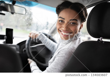 Driver turns to passenger seat and smiles at camera. Happy millennial african american woman taxi driver sitting in car holding to steering wheel, next to smartphone with blank screen, panorama 134083607
