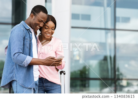 Cheerful black couple with smartphone booking hotel online after arrival to new country, romantic african american spouses standing near airport building, making accommodation reservation in internet 134083615
