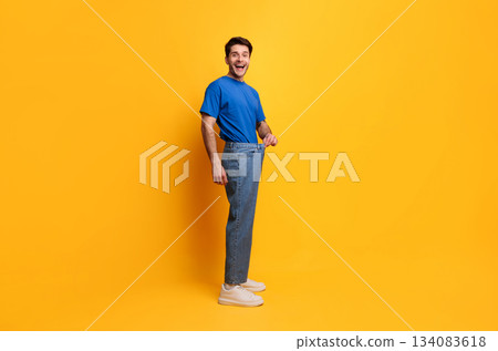 Successful Weight Loss. Happy Excited Slim Young Guy Pulling Old Large Loose Jeans Achieved Slimming Weightloss Goal Standing Posing Isolated On Yellow Studio Wall, Full Body Length, Free Copy Space Successful Weight Loss. Happy Excited Slim Young Guy Pulling Old Large Loose Jeans Achieved Slimming Weightloss Goal Standing Posing Isolated On Yellow Studio Wall, Full Body Length, Free Copy Space 134083618