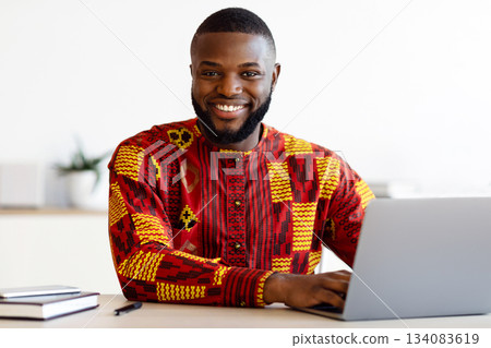 Portrait Of Smiling Black Freelancer Guy In African Traditional Clothes Sitting At Workplace In Home Office, Cheerful Man In Ethnic Costume Posing At Camera While Working With Laptop Computer Portrait Of Smiling Black Freelancer Guy In African Traditional Clothes Sitting At Workplace In Home Office, Cheerful Man In Ethnic Costume Posing At Camera While Working With Laptop Computer 134083619