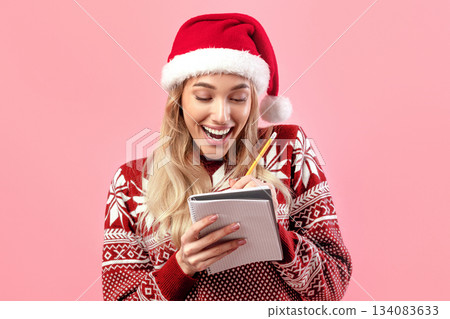 Happy blonde lady in Xmas outfit writing New Year resolution or Christmas wishlist in notebook, making plans for future on pink studio background. Millennial woman setting goals for upcoming year 134083633
