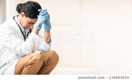 Stressed brunette young woman doctor having difficult day at clinic, wearing protective gloves, sitting next to wall in her office and resting, empty space. Upset female doctor feeling exhausted Stressed brunette young woman doctor having difficult day at clinic, wearing protective gloves, sitting next to wall in her office and resting, empty space. Upset female doctor feeling exhausted 134083634