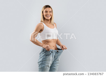 Diet, healthy lifestyle, slimming concept. Happy cheerful beautiful blonde middle aged woman wearing big jeans after weight loss on grey studio background, smiling at camera, copy space Diet, healthy lifestyle, slimming concept. Happy cheerful beautiful blonde middle aged woman wearing big jeans after weight loss on grey studio background, smiling at camera, copy space 134083767