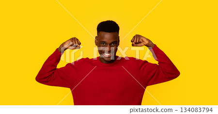 Funny Strong African American Guy In Red Sweater Showing Biceps Muscles And Looking At Camera, Cheerful Young Man Demonstrating His Strength, Standing Isolated Over Yellow Background, Copy Space 134083794