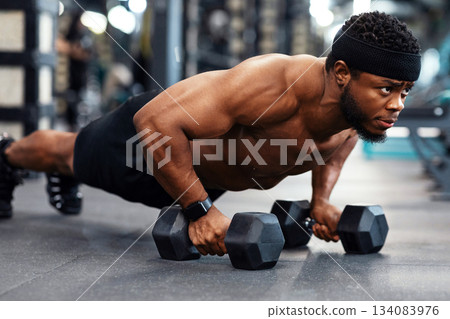 Concentrated black guy sportsman pushing up with dumbbells. Muscular african american bodybuilder shirtless man holding barbells and making push-ups from floor, work out at gym Concentrated black guy sportsman pushing up with dumbbells. Muscular african american bodybuilder shirtless man holding barbells and making push-ups from floor, work out at gym 134083976