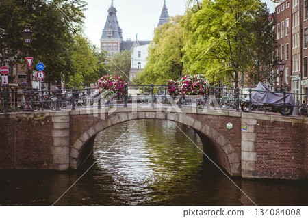 Stone arch canal bridge is spanning canal in flat style, with bicycles and hanging flower baskets 134084008