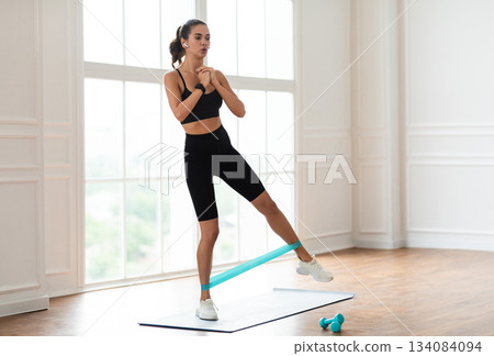 Body Shaping Workout Concept. Focused Fit Woman In Black Sportswear Exercising With Resistance Loop Band Lifting Leg Standing On Yoga Mat, Wearing Wireless Earbuds, Training Near Window With Music Body Shaping Workout Concept. Focused Fit Woman In Black Sportswear Exercising With Resistance Loop Band Lifting Leg Standing On Yoga Mat, Wearing Wireless Earbuds, Training Near Window With Music 134084094