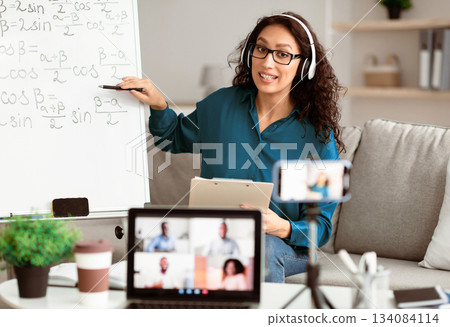 Videoconference And Training. Class of diverse students learning remotely online using web app. Female teacher in headset providing lesson looking at pc screen, recording video on cellphone on tripod 134084114