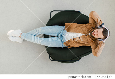 Top View Of Relaxed Guy Wearing Wireless Earphones Listening To Music Or Online Podcast Resting With Eyes Closed Lying In Chair At Home. Weekend Leisure And Relaxation Concept 134084152