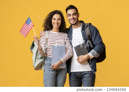Study Abroad. Smiling Middle Eastern Students Couple Posing With American Flag In Hand, Happy Young Arab Man And Woman With Backpacks And Workbooks Standing Over Yellow Studio Background, Copy Space Study Abroad. Smiling Middle Eastern Students Couple Posing With American Flag In Hand, Happy Young Arab Man And Woman With Backpacks And Workbooks Standing Over Yellow Studio Background, Copy Space 134084208