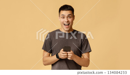 Emotions of success, victory, cashback and great message about win. Happy millennial guy student in brown t-shirt shouts with joy and holds smartphone, isolated on beige background, studio shot Emotions of success, victory, cashback and great message about win. Happy millennial guy student in brown t-shirt shouts with joy and holds smartphone, isolated on beige background, studio shot 134084233