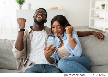 Yeah. Emotional excited black couple celebrating online win, great deal or business success, holding using smart phone, shaking clenched fists, looking at screen, guy in glasses and braces screaming 134084287