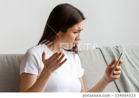 Stressed Young Pregnant Woman Looking At Smartphone Screen While Relaxing On Couch, Worried Expectant Lady Reading Unpleasant Message In Mobile Phone, Emotionally Reacting To Bad News, Free Space 134084323