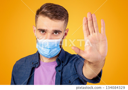 No to coronavirus. Young Caucasian man in medical mask making STOP gesture on orange studio background. Millennial guy wearing protection, expressing negative attitude to worldwide epidemic No to coronavirus. Young Caucasian man in medical mask making STOP gesture on orange studio background. Millennial guy wearing protection, expressing negative attitude to worldwide epidemic 134084329