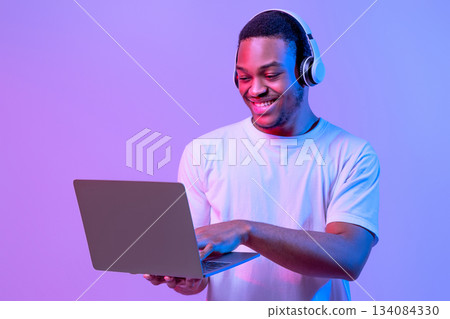 Online Gaming. Portraif Of Happy Black Man In Wireless Headphones Using Laptop Computer, Playing Games In Internet While Standing In Neon Lighting Over Purple Studio Background, Copy Space 134084330