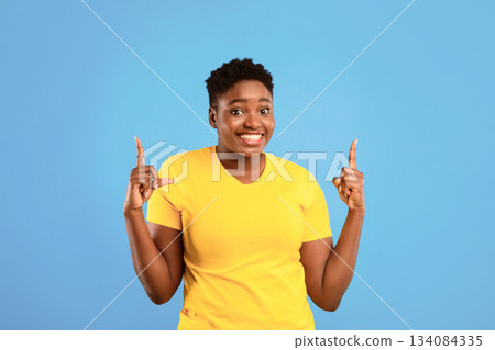 Look Upward. Cheerful African Plus-Size Lady Pointing Fingers Up Standing Posing On Blue Studio Background. Plump Black Female Showing Look There Advertising Something Upward 134084335