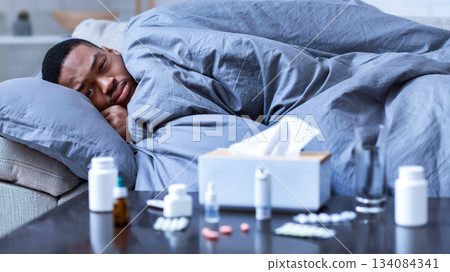 Sick Black Guy Having Fever And Chills Lying Wrapped In Blanket On Couch At Home, Looking At Camera. Covid-19 Coronavirus Disease, Seasonal Flu And Cold, Illness Fatigue. Selective Focus 134084341