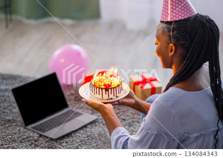 African woman in party hat celebrating birthday online at quarantine or self-isolation, using laptop with empty screen for video call with friends and family, holding cake, sitting on floor African woman in party hat celebrating birthday online at quarantine or self-isolation, using laptop with empty screen for video call with friends and family, holding cake, sitting on floor 134084343