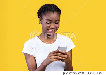 Cheerful Black Woman Using Phone Texting And Networking Standing Over Yellow Studio Background. African Young Lady Using Mobile Application On Smartphone. Empty Space For Text 134084350