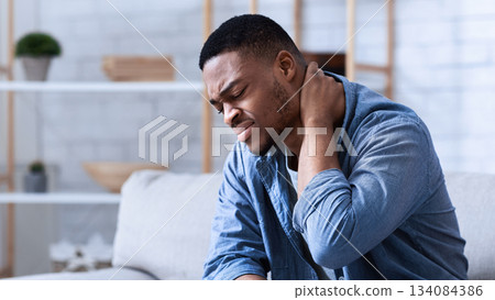 Black Man Suffering From Neck Pain Massaging Aching Muscles Sitting On Couch In Living Room At Home. Neurological Health Problem, Arthritis And Osteoarthritis Concept Black Man Suffering From Neck Pain Massaging Aching Muscles Sitting On Couch In Living Room At Home. Neurological Health Problem, Arthritis And Osteoarthritis Concept 134084386