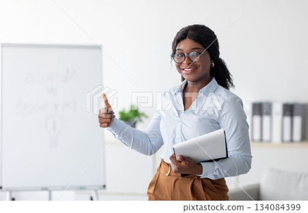 Psychology teacher or business coach giving seminar or presentation, showing thumb up gesture at office. Black female lecturer recommending training course on personal growth and motivation Psychology teacher or business coach giving seminar or presentation, showing thumb up gesture at office. Black female lecturer recommending training course on personal growth and motivation 134084399
