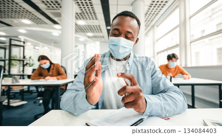 Hygiene And Protection Concept. African american guy applying antibacterial sanitizer spray on hands, wearing surgical face mask, sitting in classroom. Prevent the spread of infection and germs 134084403