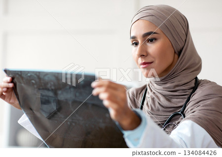 Portrait of muslim arab female medical doctor looking at xray scan, clinic interior, copy space. Young woman in hijab physician holding CT scan, checking disease progress or treatment efficiency Portrait of muslim arab female medical doctor looking at xray scan, clinic interior, copy space. Young woman in hijab physician holding CT scan, checking disease progress or treatment efficiency 134084426