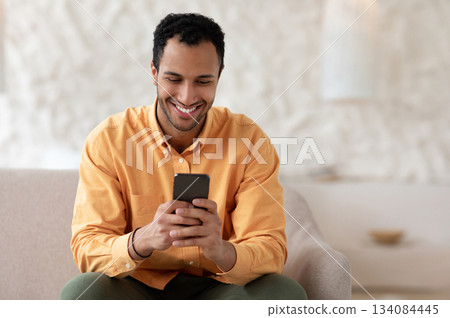 Cool Gadget And Application. Portrait of young smiling Arab man holding mobile phone, typing sms message, sitting on the couch in living room. Guy browsing internet, surfing web, using app, free space Cool Gadget And Application. Portrait of young smiling Arab man holding mobile phone, typing sms message, sitting on the couch in living room. Guy browsing internet, surfing web, using app, free space 134084445