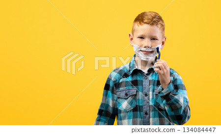 Playful little young boy shaving face over yellow studio background, imitating his dad, copy space. Cute redhead kid with shaving foam on his face holding razor, acting like adult man Playful little young boy shaving face over yellow studio background, imitating his dad, copy space. Cute redhead kid with shaving foam on his face holding razor, acting like adult man 134084447