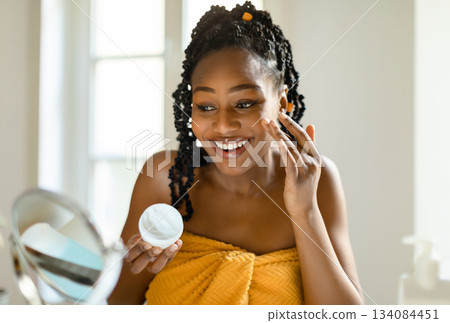 Daily beauty routine. Happy black lady applying moisturising cream on face, looking at magnifying mirror in bedroom, using nourishing skin and smiling, free space 134084451