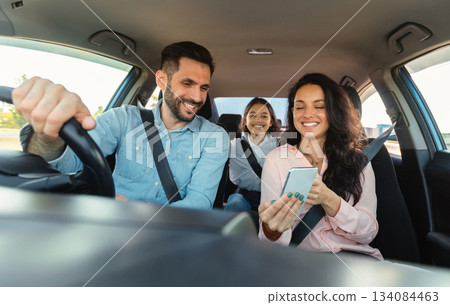 Car navigation app. Happy woman showing gps application on cellphone to driver man, family travelling and navigating vehicle with gadget, enjoying weekend trip Car navigation app. Happy woman showing gps application on cellphone to driver man, family travelling and navigating vehicle with gadget, enjoying weekend trip 134084463