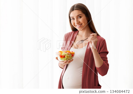 Nutrition During Pregnancy. Smiling Pregnant Lady Eating Fresh Vegetable Salad Near Window, Holding Bowl With Vegetarian Meal And Looking At Camera, Enjoying Healthy Food While Expecting Baby Nutrition During Pregnancy. Smiling Pregnant Lady Eating Fresh Vegetable Salad Near Window, Holding Bowl With Vegetarian Meal And Looking At Camera, Enjoying Healthy Food While Expecting Baby 134084472