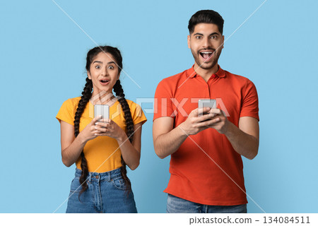Mobile Offer. Portrait Of Happy Shocked Arab Couple Holding Smartphones In Hands And Looking At Camera With Excitement, Young Middle Eastern Man And Woman Opening Mouth With Amazement, Copy Space 134084511