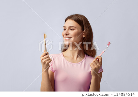 Smiling young woman holding eco bamboo and plastic toothbrush, choosing between different items for dental hygiene. Eco-friendly lady looking at wooden brush, standing on grey background, copy space 134084514