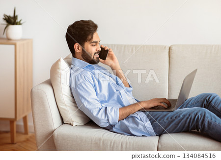 Freelance Concept. Handsome Arab Man Using Laptop And Talking On Cellphone At Home, Millennial Eastern Guy Working Remotely While Sitting On Couch In Living Room, Enjoyin Distant Job, Side View 134084516