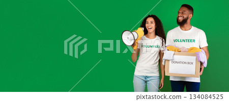 Volunteering And Charity. Female And Male Volunteers Holding Donation Box Collecting Clothes For Homeless People Standing Over Green Studio Background. Panorama With Copy Space Volunteering And Charity. Female And Male Volunteers Holding Donation Box Collecting Clothes For Homeless People Standing Over Green Studio Background. Panorama With Copy Space 134084525