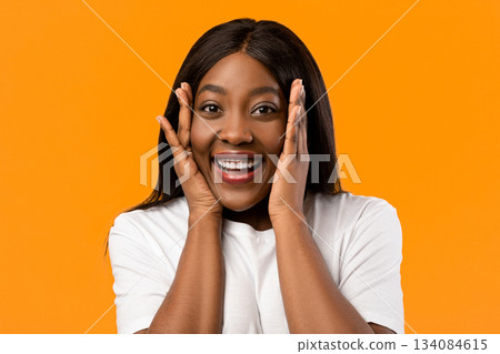 Closeup of emotional black lady in white t-shirt smiling and touching her face over orange studio background, emotional african american young woman expressing amazement, seeing something exciting Closeup of emotional black lady in white t-shirt smiling and touching her face over orange studio background, emotional african american young woman expressing amazement, seeing something exciting 134084615
