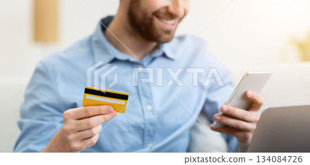 Joyful guy shopping online with cellphone and credit card sitting on couch at home, focus on bank card. Mobile shopping application for phone, e-commerce, banking app concept. Shallow depth 134084726
