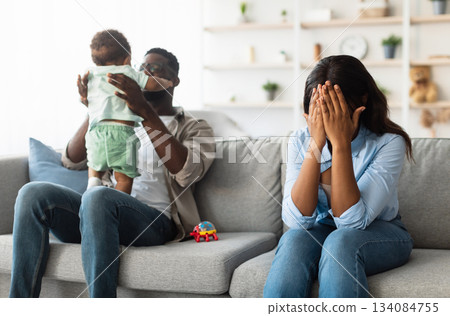 Post Partum Depression. Exhausted black mom covering face crying sitting on couch at home, feeling headache and stress, her husband playing with small infant kid. Challenges of Parenthood. Baby blues 134084755