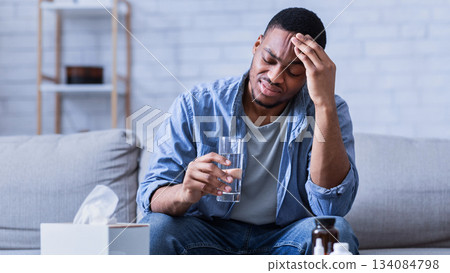 Unhappy Black Guy Suffering From Headache Or Migraine Holding Glass Of Water Taking Pills Sitting On Sofa At Home. Painkilling Medicine, Medical Treatment Concept. Selective Focus 134084798
