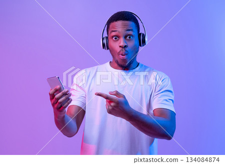 Cool App. Surprised Black Guy In Wireless Headphones Pointing At Smartphone In Hand, Emotionally Reacting To New Mobile Application Or Music Streaming Service, Standing Under Neon Light, Copy Space 134084874
