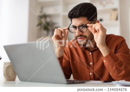 Error. Portrait of confused Arab guy sitting at desk using laptop, taking off glasses looking at pc screen and squinting. Worried man reading bad negative news, having poor eyesight problems 134084876